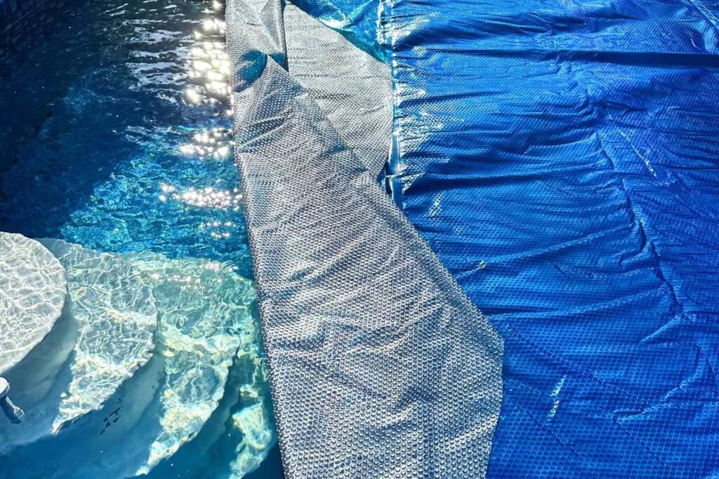 A blue solar pool cover floating on a backyard swimming pool.