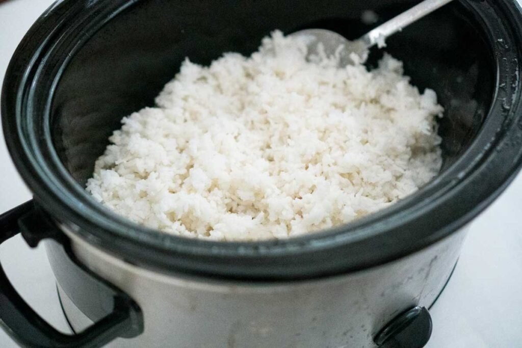 A bowl of fluffy cooked rice sits beside a slow cooker in a kitchen.