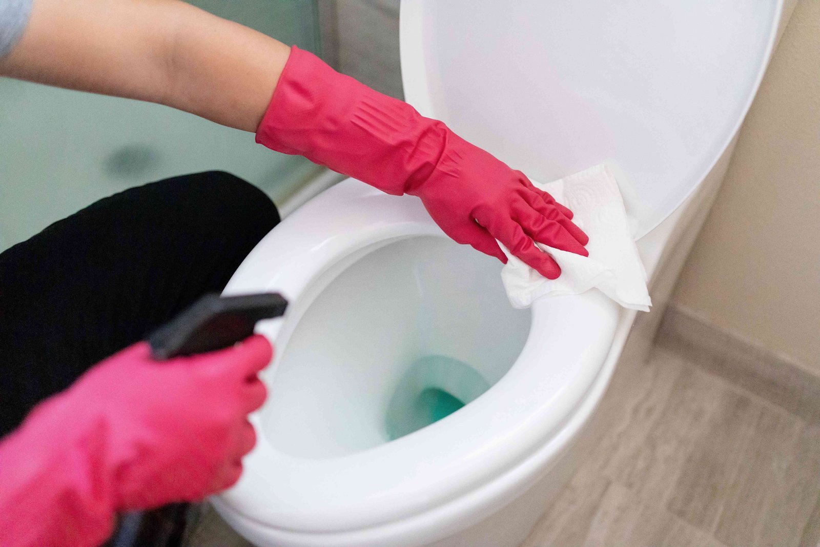 Hands wearing rubber gloves preparing to clean a white toilet seat cover in a bathroom.