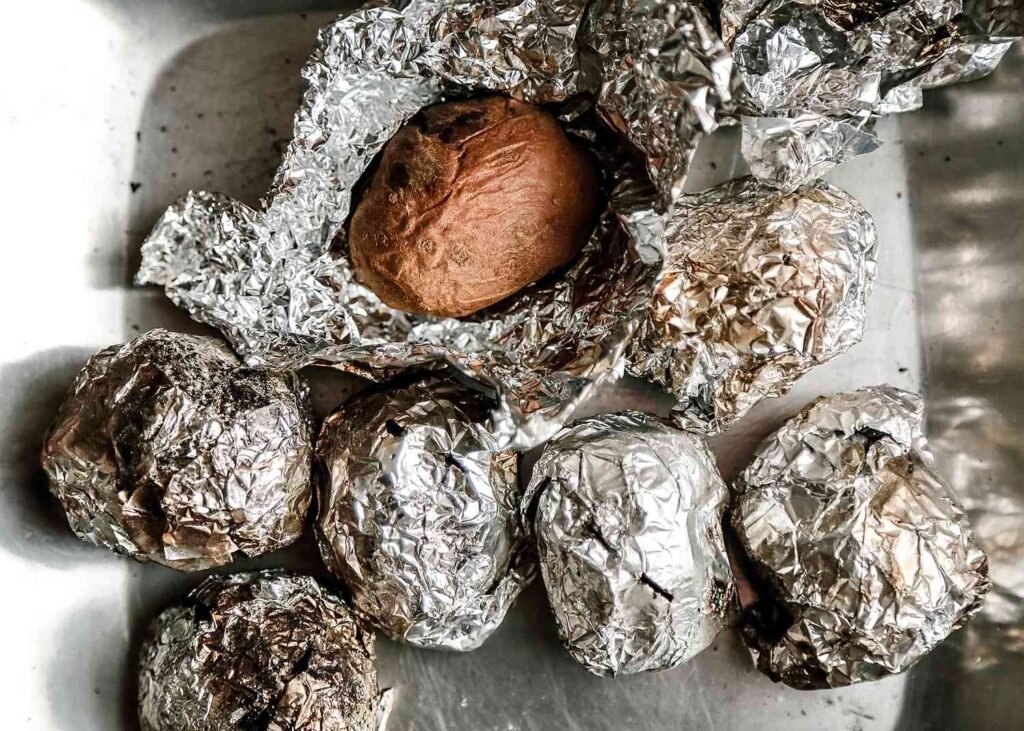 A variety of baked potatoes wrapped in foil and unwrapped on a wooden table.