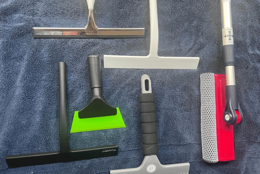 A hand using a handheld silicone squeegee to clean a clear glass shower door.