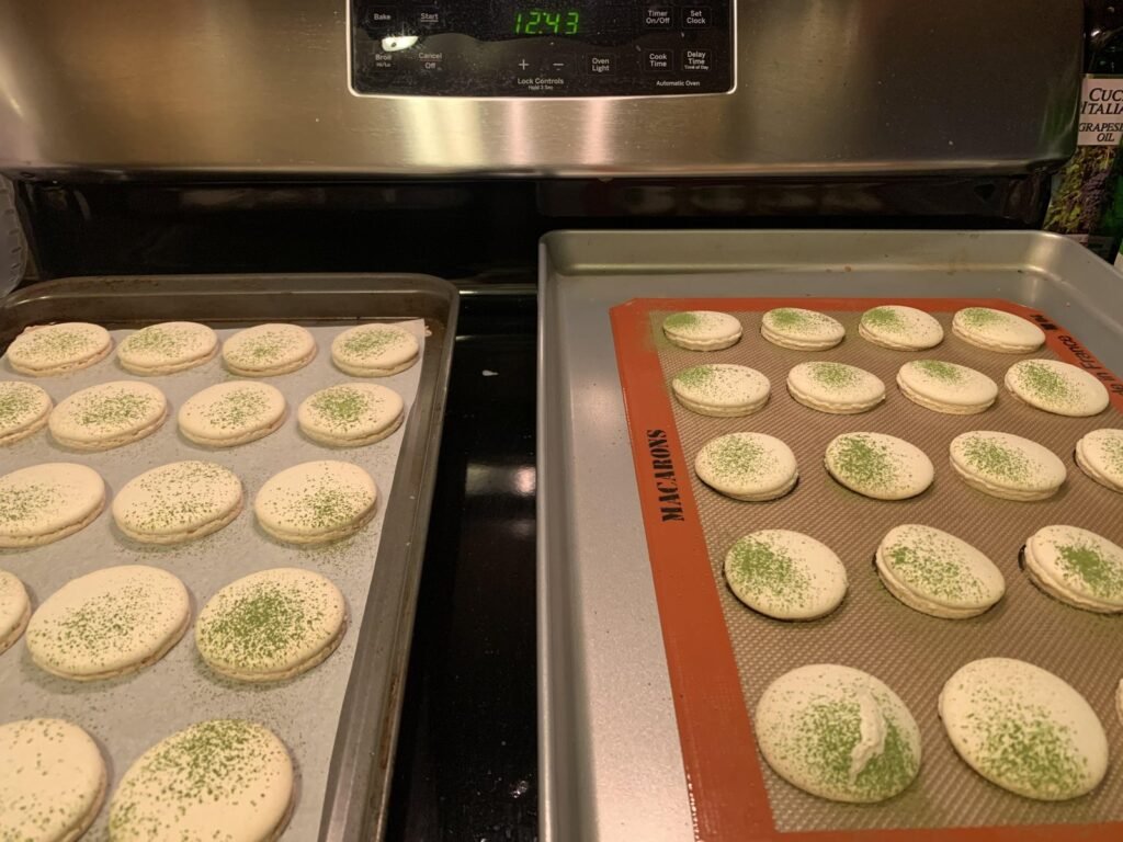Macaron shells piped on a dark nonstick Teflon baking sheet on a marble counter